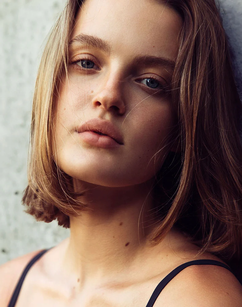 Close-up portrait of a woman with a bob haircut and blue eyes leaning against a wall with a neutral expression.