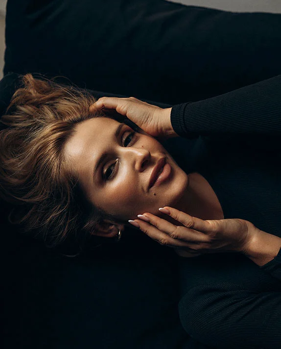 A woman lies back with her hands in her hair, looking into the camera with a radiant and rejuvenated facial appearance.