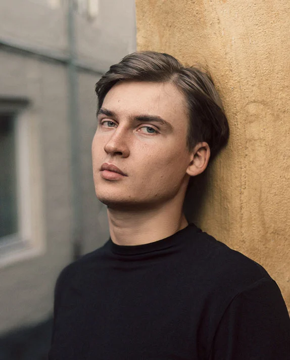 A young man with short brown hair leans against a textured tan wall, presented in a clean profile that emphasizes refined earlobe aesthetics.