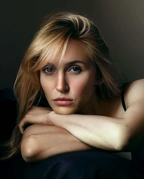 A close-up, high-fashion portrait of a woman with blonde hair resting her chin on her crossed arms, showcasing a smooth jawline and youthful facial contours.