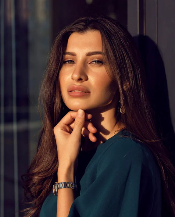 A woman with long brown hair looks into the distance while resting her hand on her chin, captured in warm, natural light that highlights youthful facial volume.