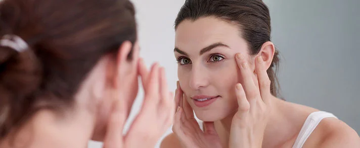A woman looking in a mirror and gently touching the skin around her eyes, examining her complexion.