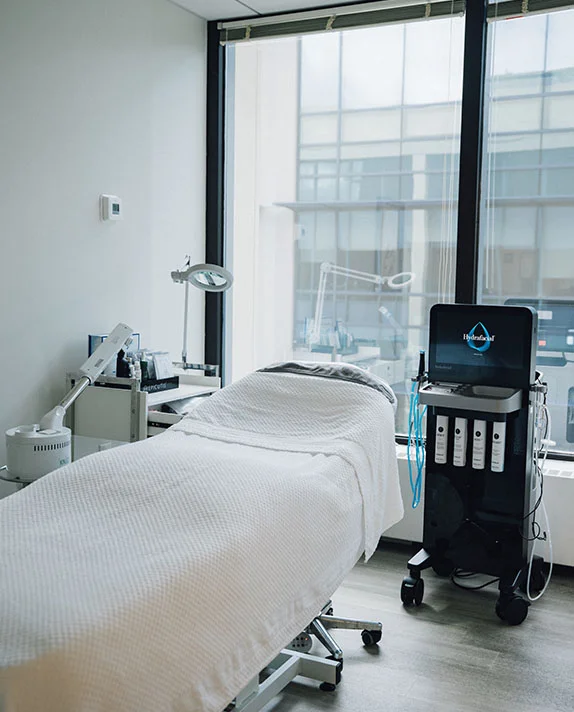 A clean, bright medical treatment room featuring a white upholstered procedure bed and a HydraFacial machine positioned near a large window.