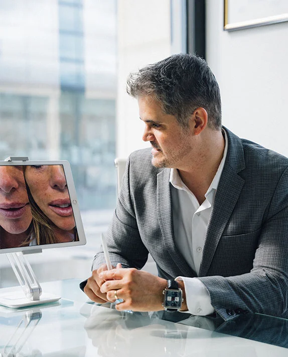 Dr. Abraham Pathak conducting a patient consultation, pointing to a digital screen showing side-by-side facial before-and-after results to a female patient.