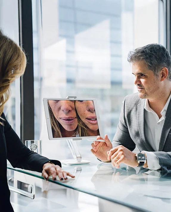 Dr. Abraham Pathak conducting a patient consultation, pointing to a digital screen showing side-by-side facial before-and-after results to a female patient.
