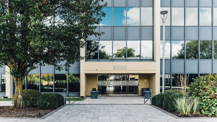 The exterior entrance of Dr. Abraham Pathak’s office at building 3020, featuring a clean, modern glass facade and a paved walkway lined with manicured greenery.