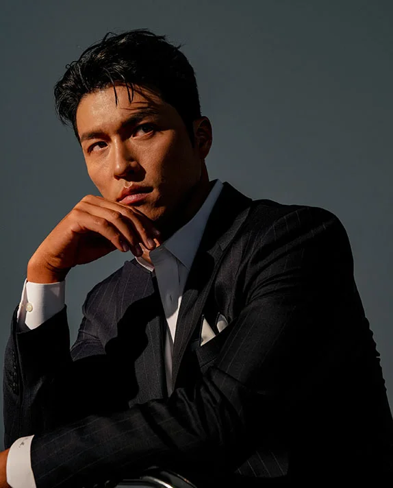 A man in a dark pinstripe suit and white dress shirt poses with his hand on his chin, showcasing a sharp and masculine jawline in high-contrast lighting.