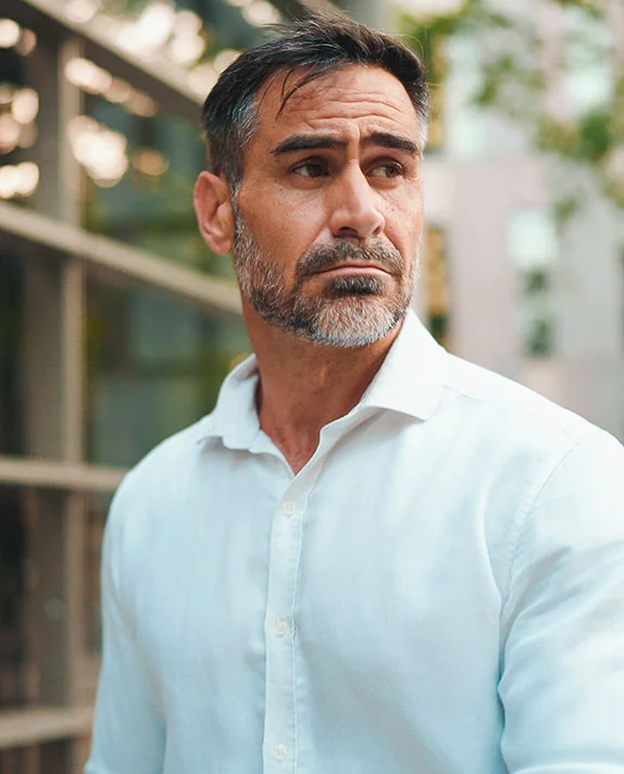 A portrait of a mature man with a groomed grey beard and white shirt looking thoughtfully to the side, highlighting the natural-looking results of a male blepharoplasty.