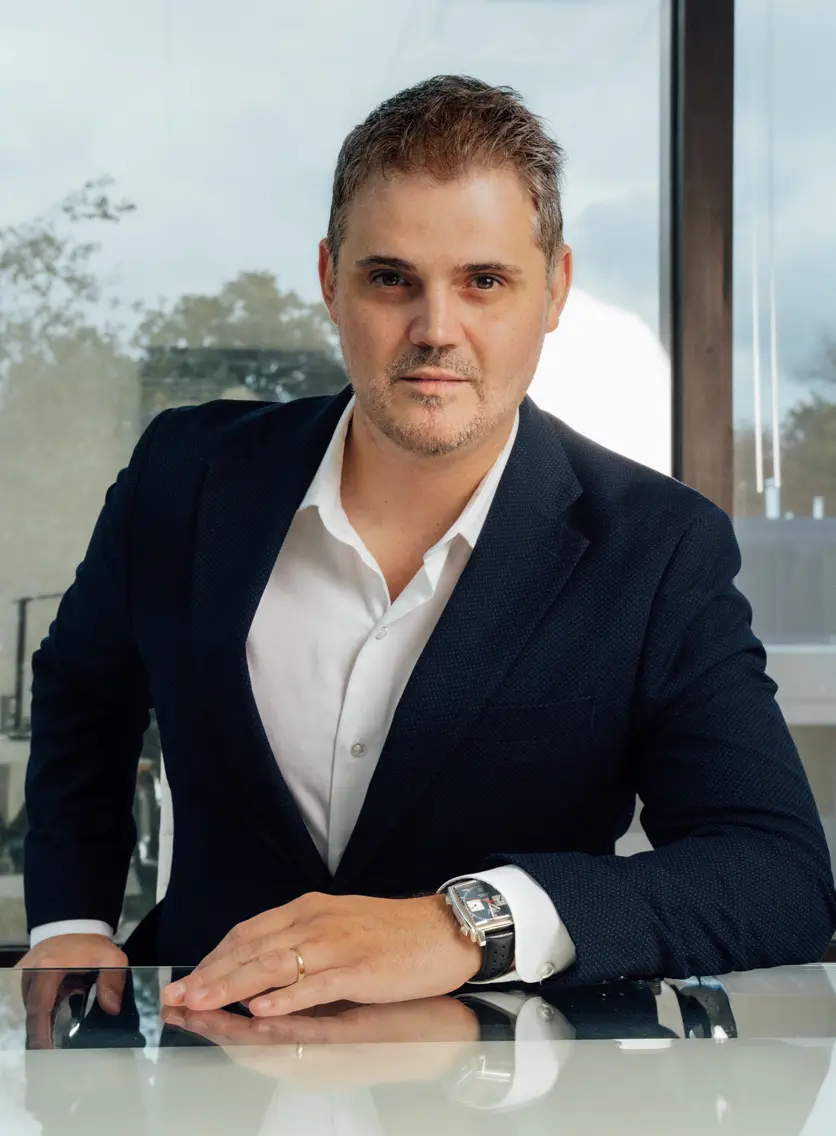 A professional portrait of Dr. Abraham Pathak, a plastic surgeon, leaning forward at a reflective desk in a modern office with large windows in the background.