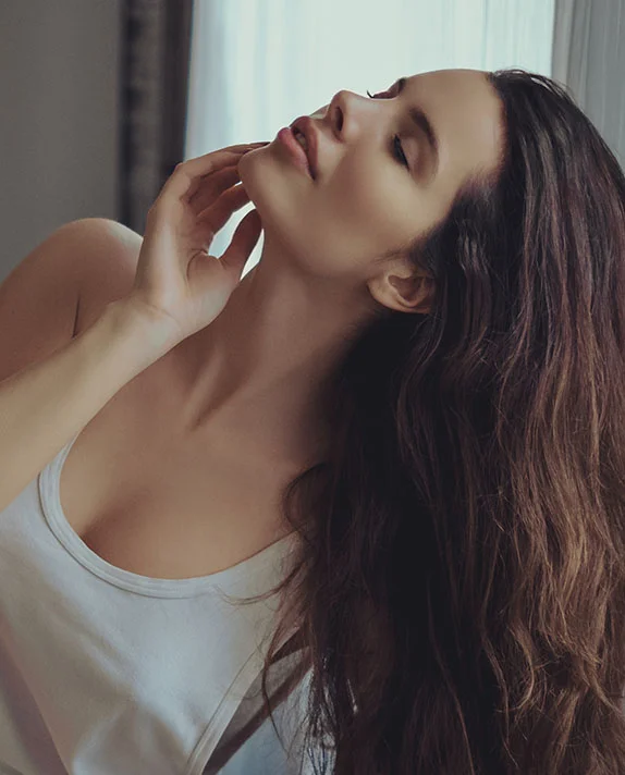 A woman with long, wavy dark hair tilts her head back and gazes upward, resting her hand gently against her jawline to showcase her neck and profile.