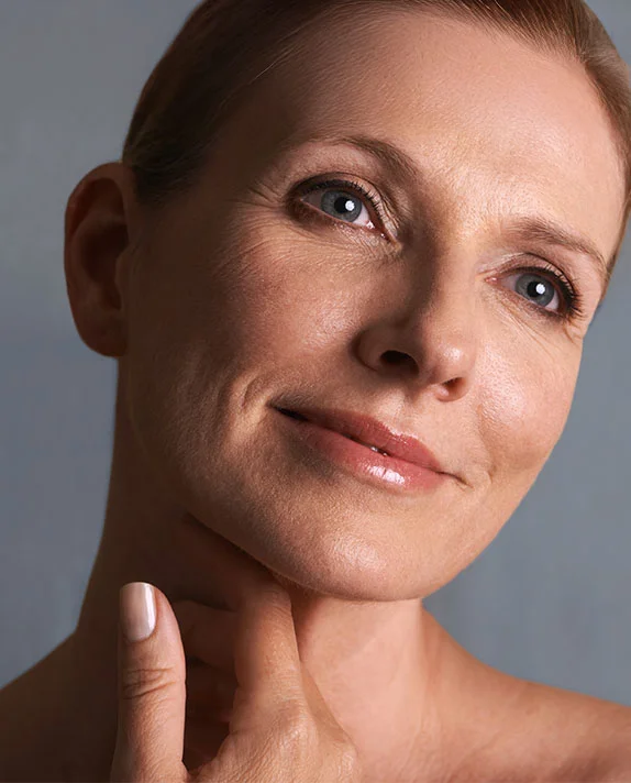 A close-up, slightly angled portrait of a mature woman with soft skin, looking upward with a gentle smile while her hand rests lightly on her neck.