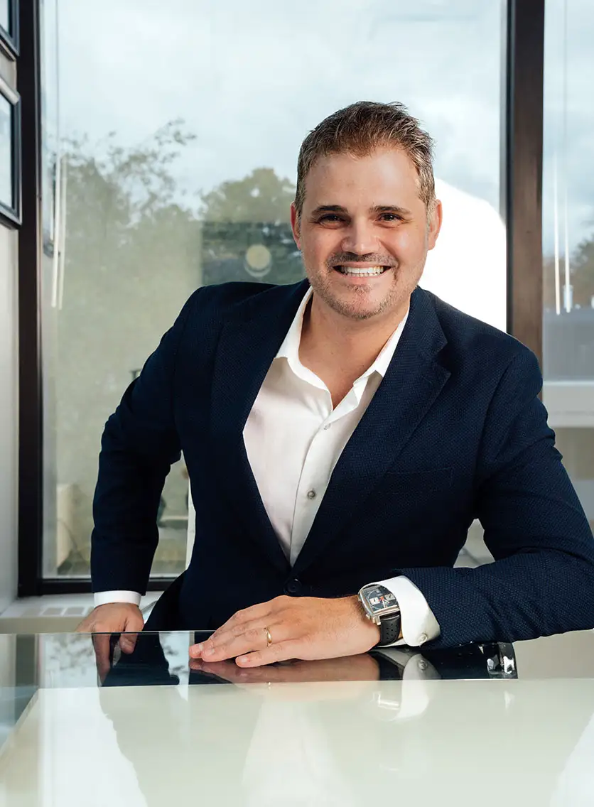 A professional portrait of Dr. Abraham Pathak, a plastic surgeon, leaning forward at a reflective desk in a modern office, smiling, with large windows in the background.