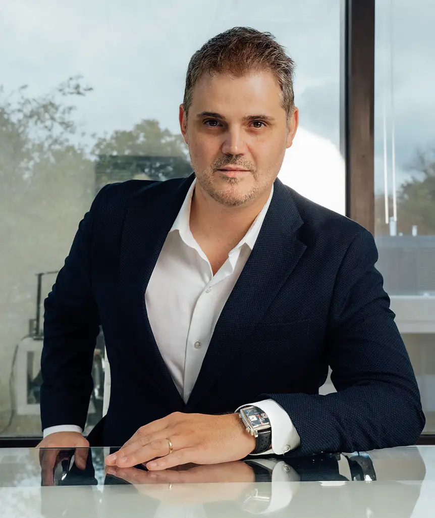 A professional portrait of Dr. Abraham Pathak, a plastic surgeon, leaning forward at a reflective desk in a modern office with large windows in the background.