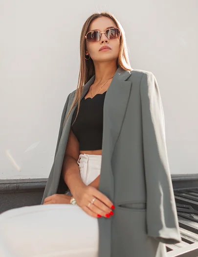 A fashionable woman wearing sunglasses, a black crop top, and a draped green blazer, embodying the confidence and lifestyle of a post-procedure patient.