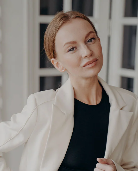 A woman with red-toned hair pulled back into a sleek bun, wearing a black top and a cream-colored blazer, looking directly at the camera with a professional expression.