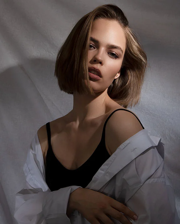A fashion-forward portrait of a woman with a sleek bob haircut wearing an open white button-down shirt over a black camisole, posed in dramatic lighting.