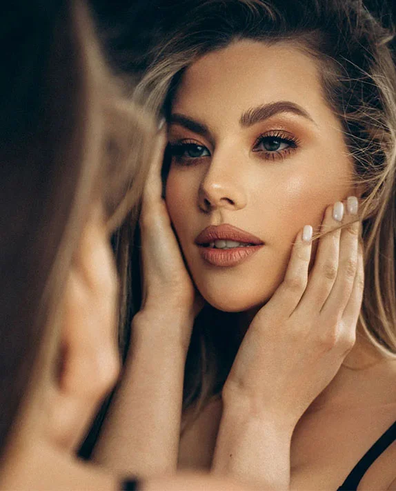 A close-up of a woman with manicured nails framing her face as she looks into a mirror, highlighting her refined facial features and profile.