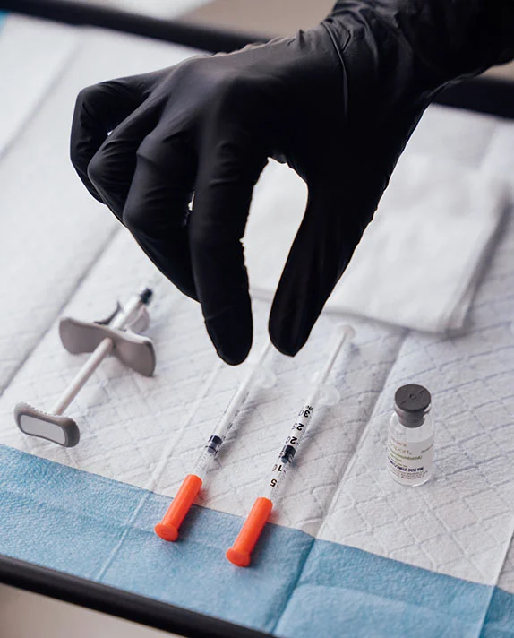 A close-up shot of a practitioner's hand in a black sterile glove reaching for syringes and a vial on a sterile medical tray, representing injectable treatments like Sculptra.