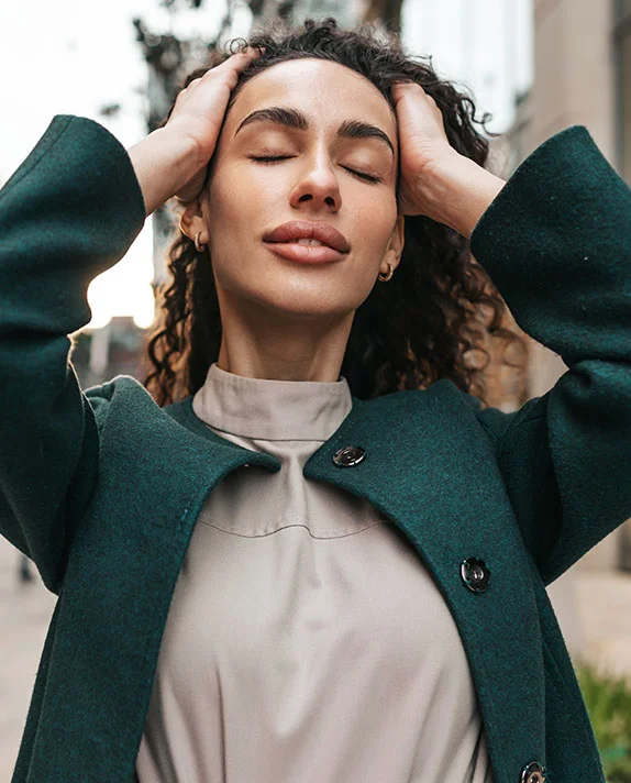 A woman with dark curly hair and closed eyes rests her hands on the sides of her head while wearing a dark green coat.